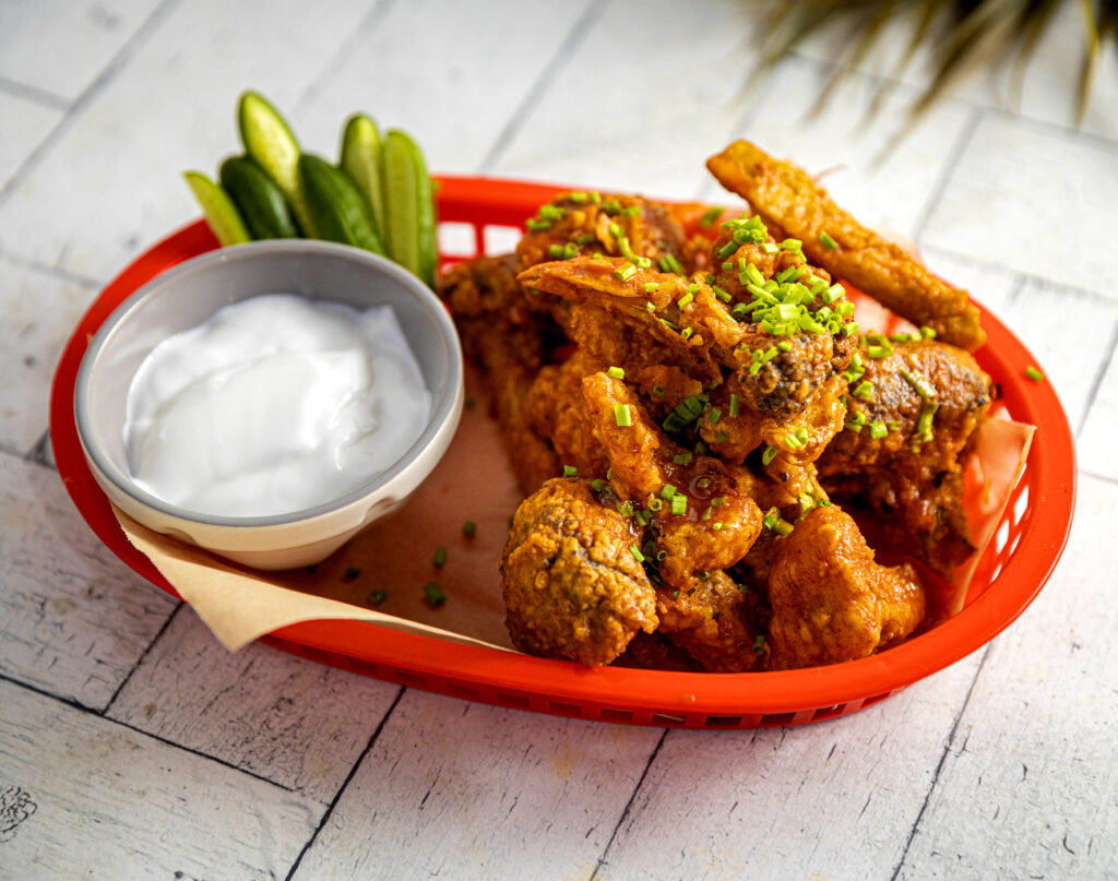 A diner-style bowl is filled with crumbed and fried broccoli and cauliflower bites, with a side of a creamy sauce and crunchy pickles.