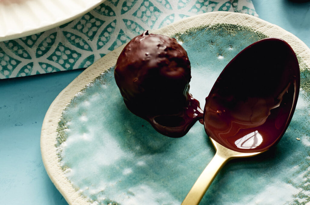 A close up image of a chocolate covered truffle on a plate with a teaspoon covered in melted chocolate sitting beside it.