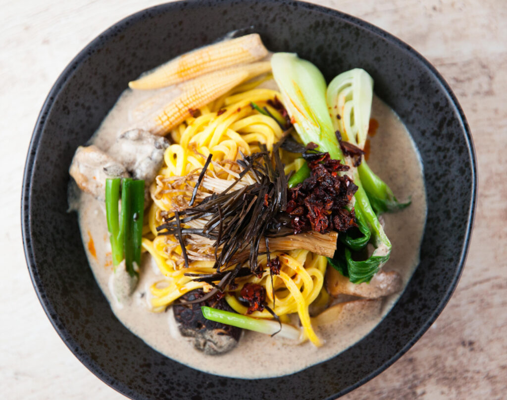 An overhead shot of ramen noodles in a bowl in a creamy broth, served with baby corn and Asian greens.