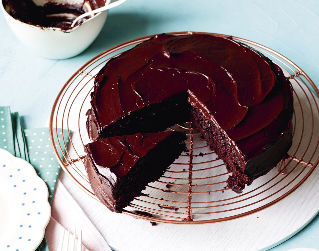 A rich chocolate cake sits on a cooling tray, covered in the most drool-worthy looking ganache.