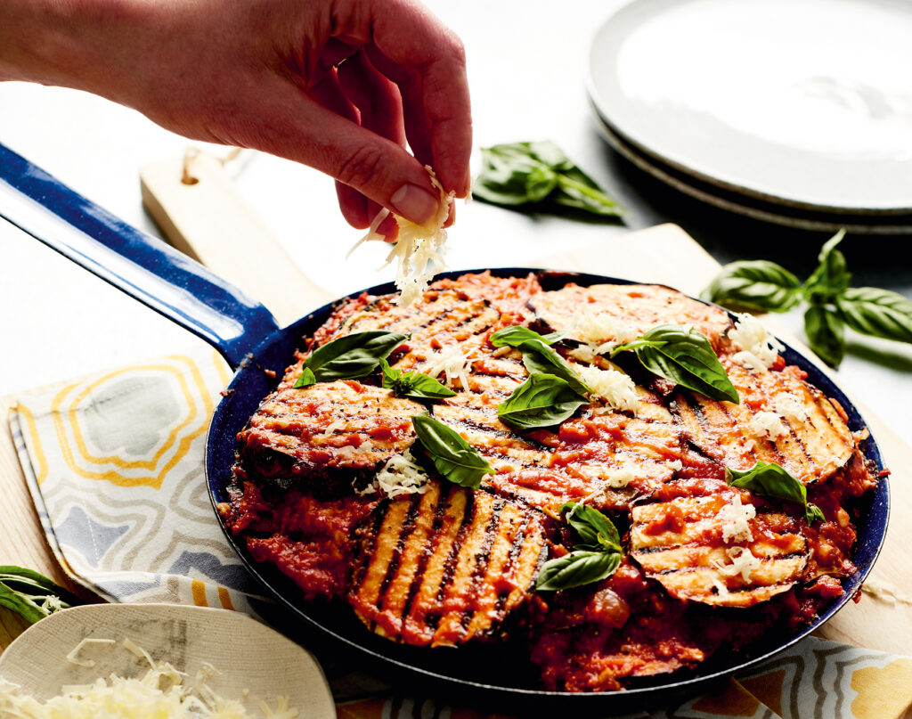 A rich and saucy eggplant bake sits in a skillet with a hand in shot topping the meal with vegan cheese.