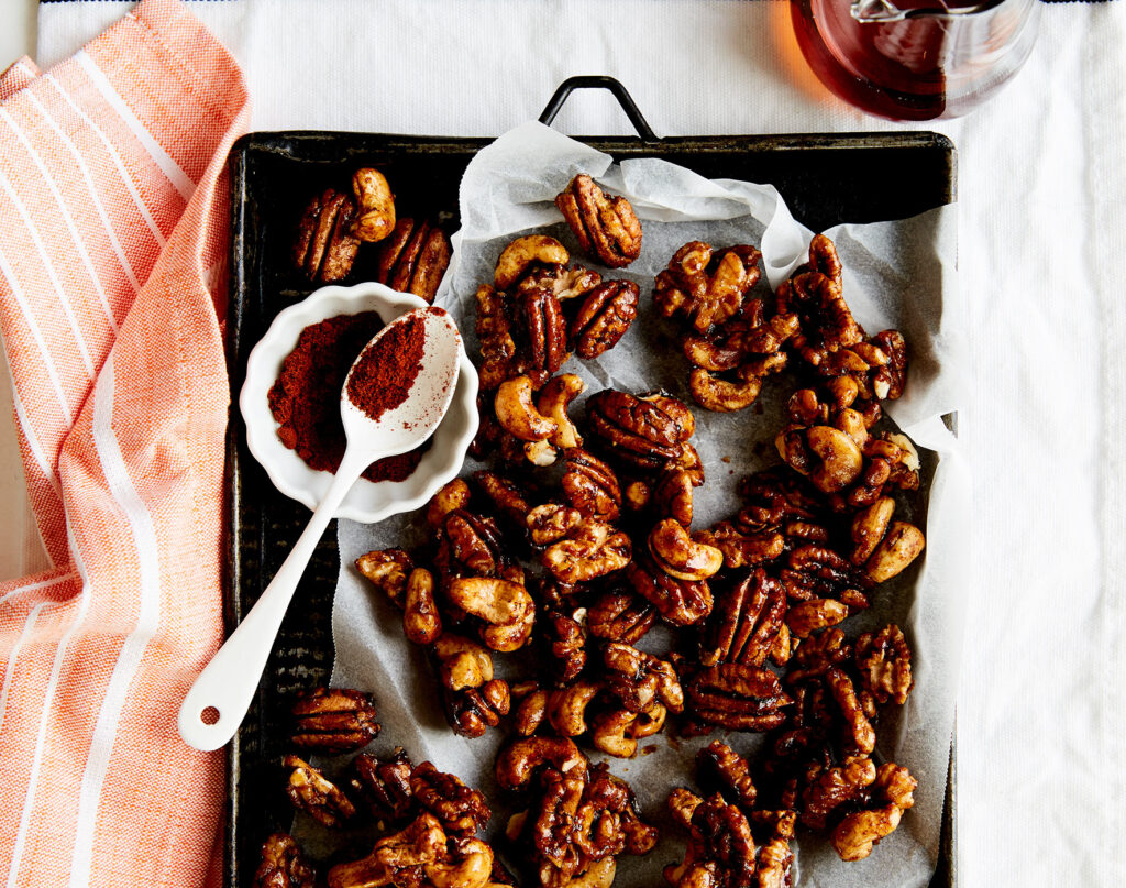 An overhead shot of roasted nuts on a baking tray lined with paper. A spoon of chilli powder sits to the side.