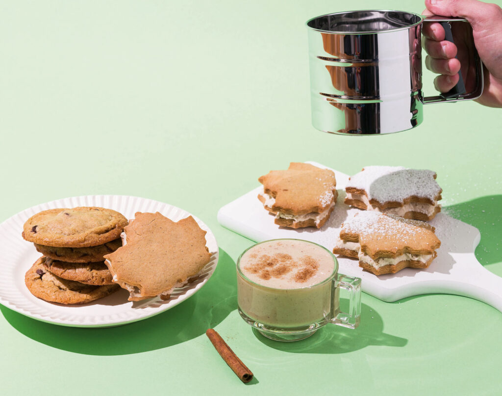 A creamy looking clear glass of eggnog on a green background. Plates of cookies sit to either side and a hand is holding a sifter above the cookies, dusting them with icing sugar. 