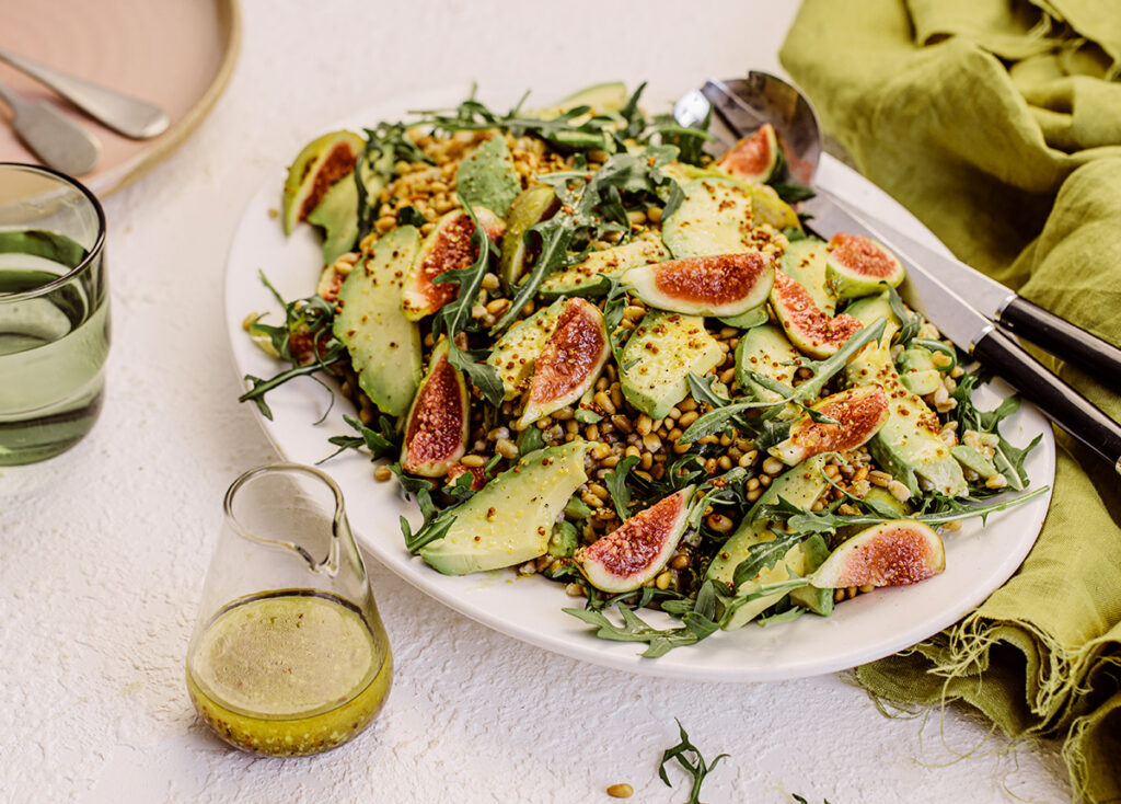 Fig, Spelt & Rocket Salad on a plate at a table setting, with a pourer of dressing on the side.