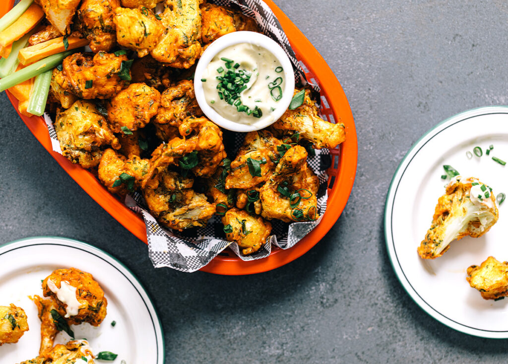 Overhead image of crispy, tempura-style cauliflower in a bowl, with several plates in shot. 