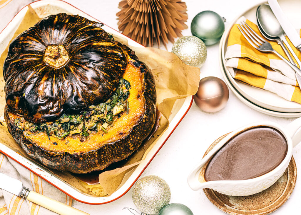 A whole roasted stuffed pumpkin sits on a tray on a white table, with a rich gravy in a boat to the side. Christmas baubles decorate the table.