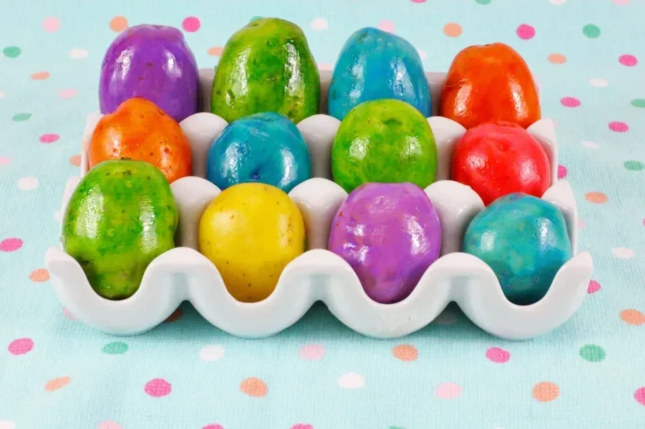 A cardboard egg tray sits on a blue polka dot fabric, filled with a dozen brightly dyed potatoes.