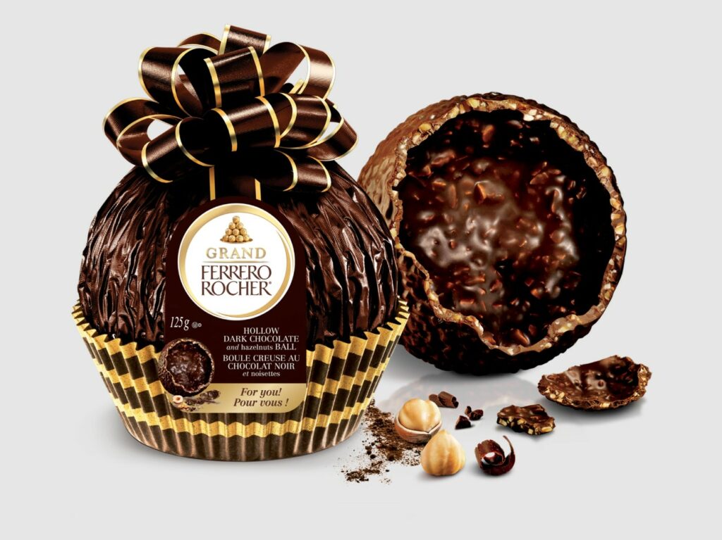 A giant dark chocolate Ferrero Rocher beautifully wrapped with an exposed and broken egg in the background.