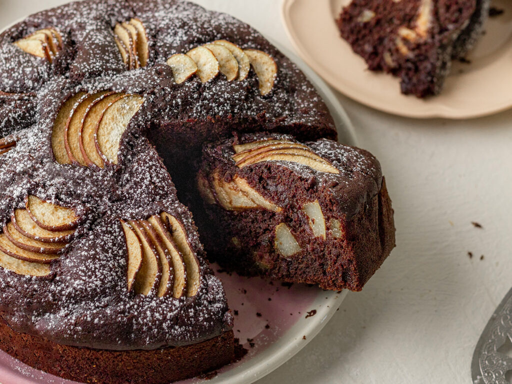 A close up image of a chocolate cake with a slice cut to show delicious pieces of pear throughout.