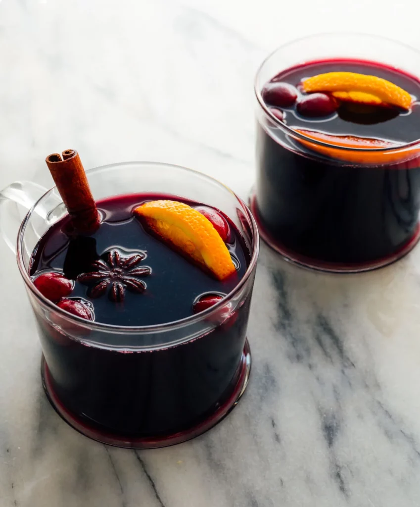 Two tumblers of mulled wine on a marbled benchtop, garnished with a cinnamon stick, cranberries, star anise and orange slices.