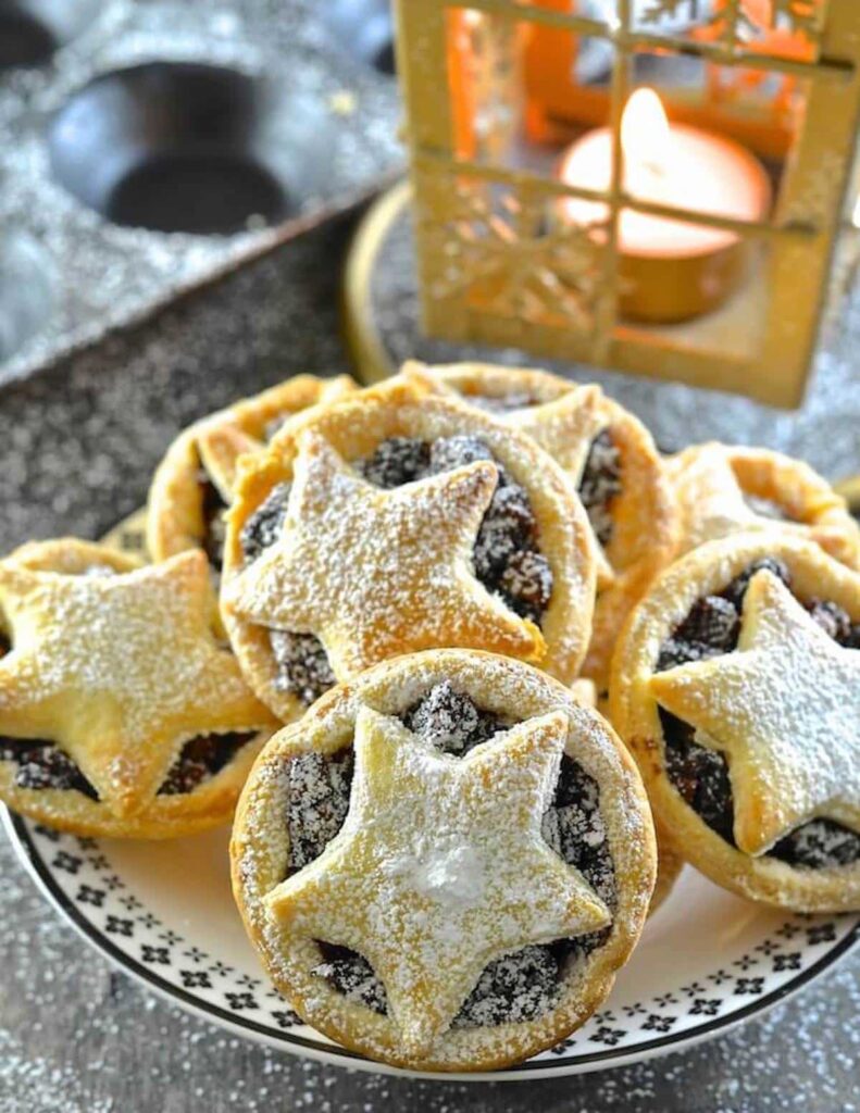 A close up image of fruit mince pies with pastry star decorations on top. A Christmas lantern is in the background with a lit tealight.