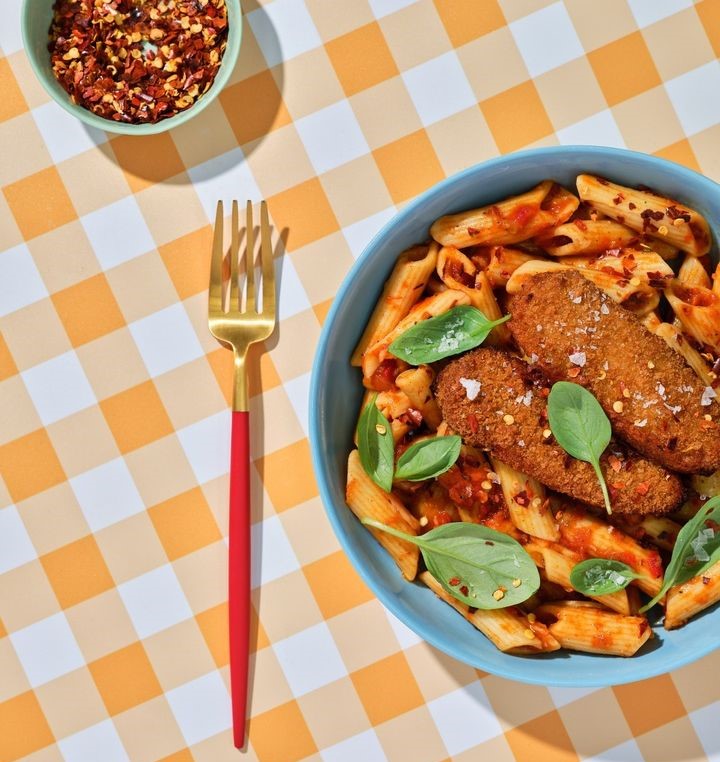 Overhead image of a pasta dish topped with roasted chicken tenders on a colourful orange checked tablecloth.