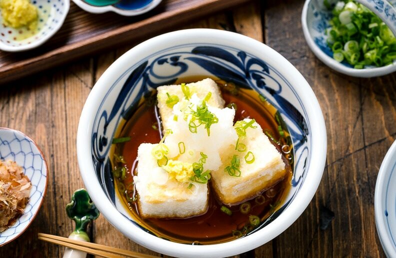 An overhead image of tofu in a small bowl with umami sauce, surrounded by small bowls with toppings and condiments.