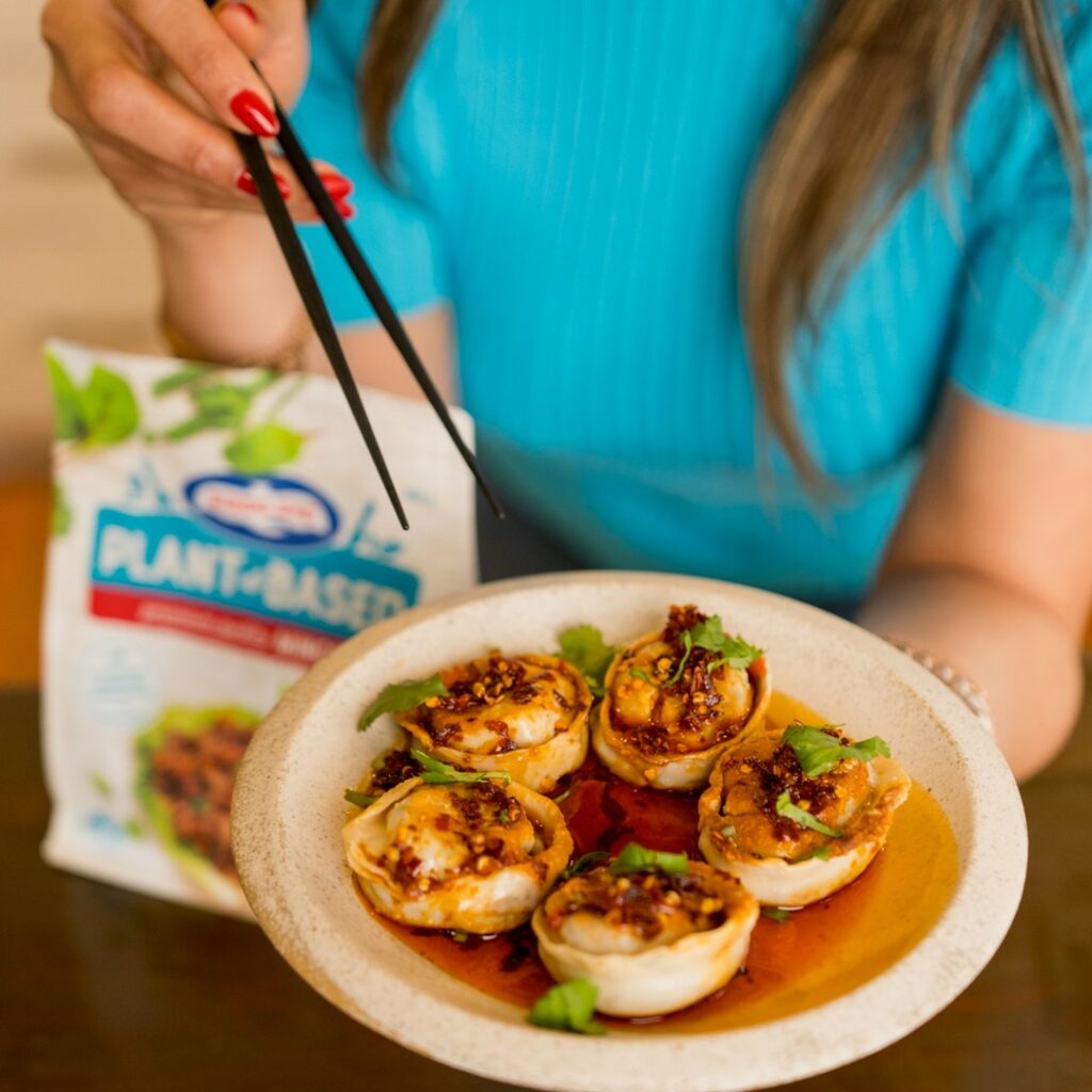 Overhead cropped shot of holding a plate of dumplings to the camera, with a plant-based mince pack in the background.