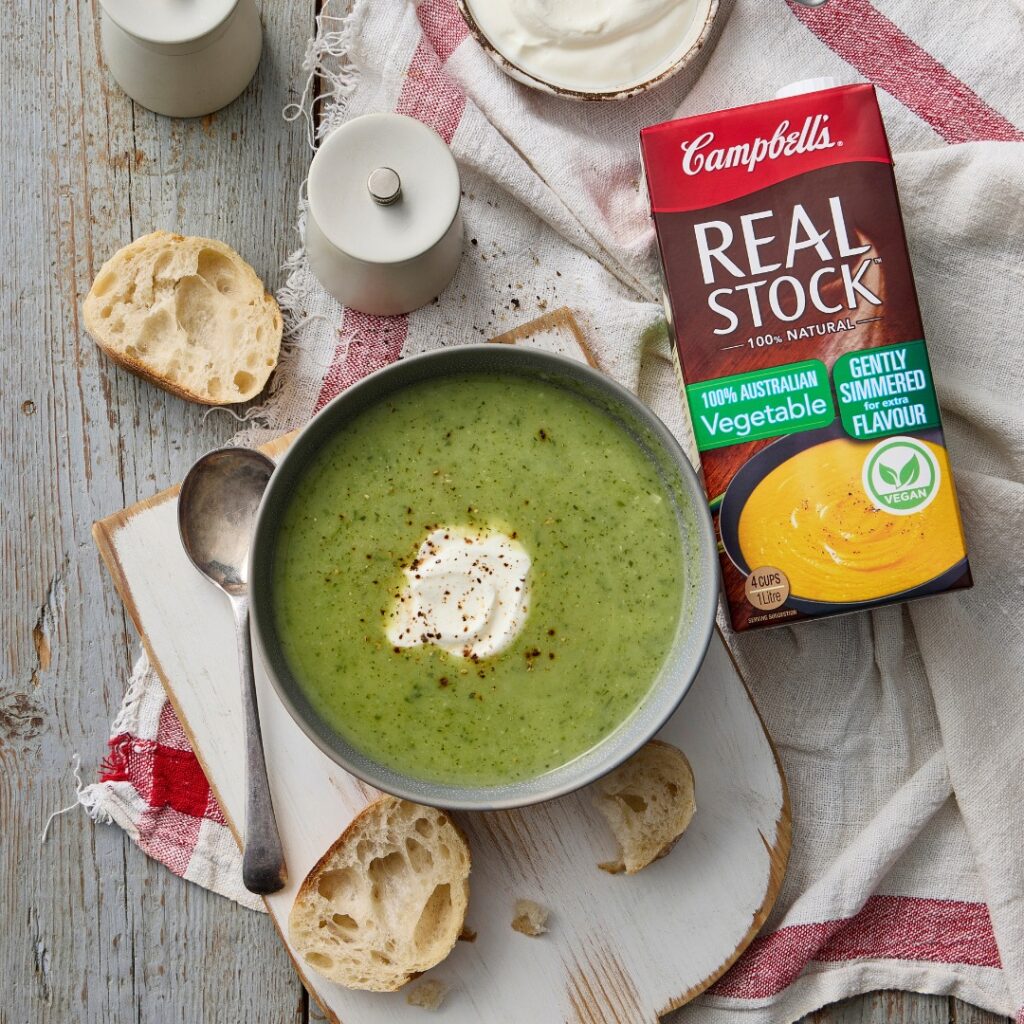 An overhead image of a green soup on a board with crusty bread, a carton of Campbell's vegetable stock sits to the side.