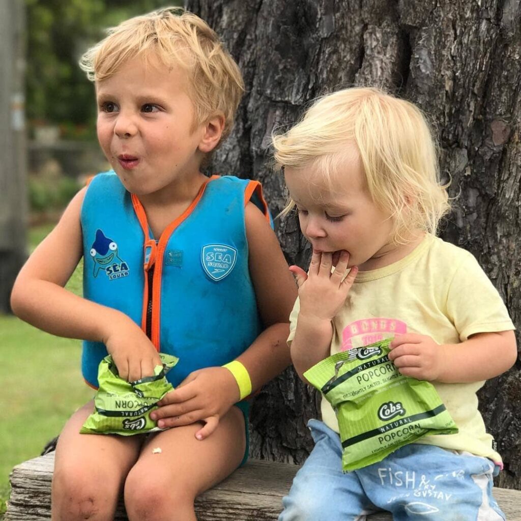 A pair of kids sit outside on a bench at the base of a tree, snacking on Cobs snack size packets of popcorn.