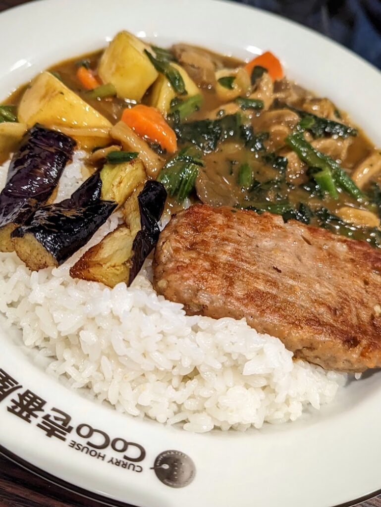 A bowl containing a bed of sticky white rice topped with grilled eggplant and a vegan soy meat patty. Beside the rice is a rich, golden-brown Japanese curry full of spinach, mushrooms, potato, and carrots. The rim of the bowl features the words 'Curry House CoCo'.