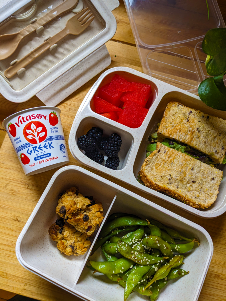 Overhead shot of an open lunchbox on a wooden bench showing variety, with a store-bought strawberry soy yoghurt to the side.