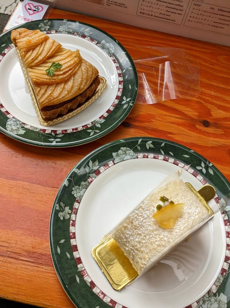 Two quaint-looking plates on a wooden cafe table. On one plate is an elaborately patterned slice of apple pie, and on the other sits a lemon cream roll topped with powdered sugar and candied lemon.