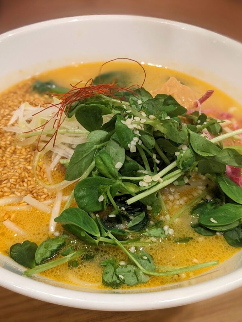 A bowl of ramen noodles in a rich golden sesame broth, garnished with pea sprouts, bean shoots, saffron, and sesame seeds.