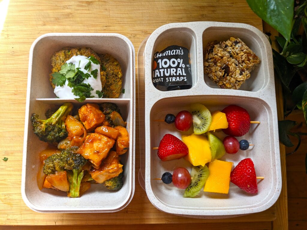 Two bento boxes sit atop a wooden bench filled with pineapple tofu, fritters, fruit skewers, cookies, and a packaged fruit strap.