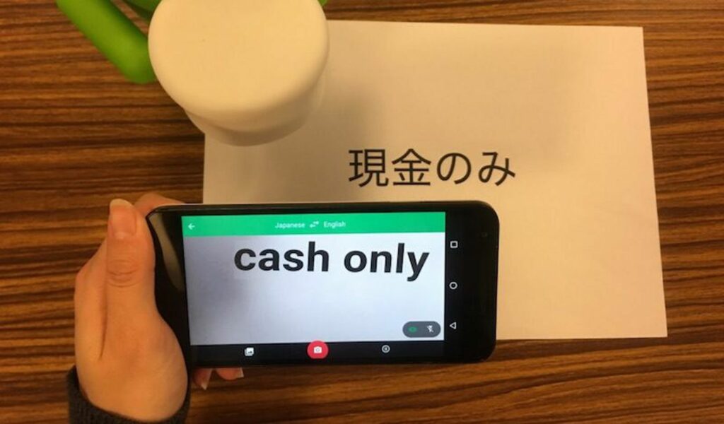 A hand holds their phone over a sign in Japanese, using Live Google Translate to show it reads 'Cash Only'.