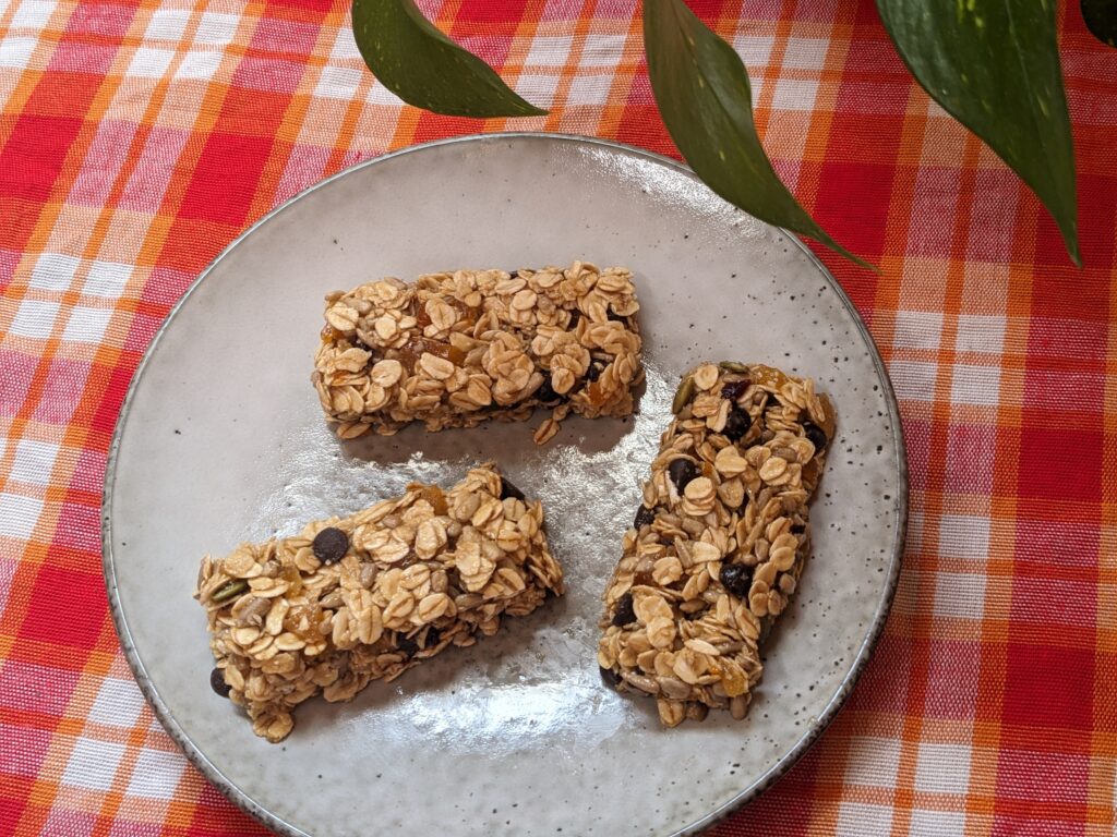 Overhead image of a plate with three rectangular granola bars. There is a plaid red, pink and orange tablecloth underneath. 