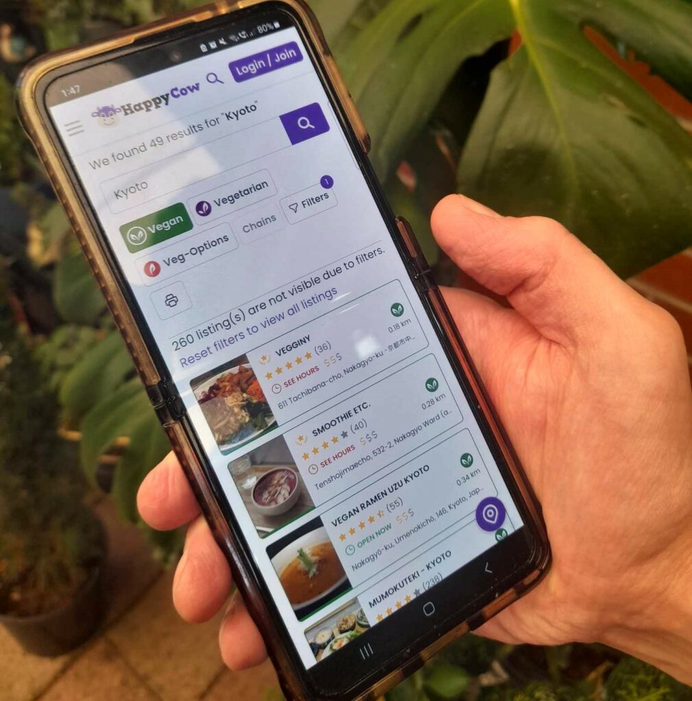 A hand holding a phone which shows a Happy Cow search for vegan restaurants and cafes in Kyoto.