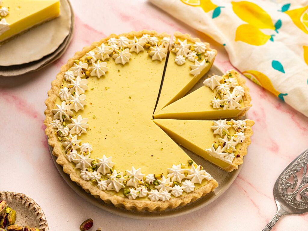 Overhead image of a creamy lemon tart, with several portions cut. It is beautifully decorated with cream and pistachios.