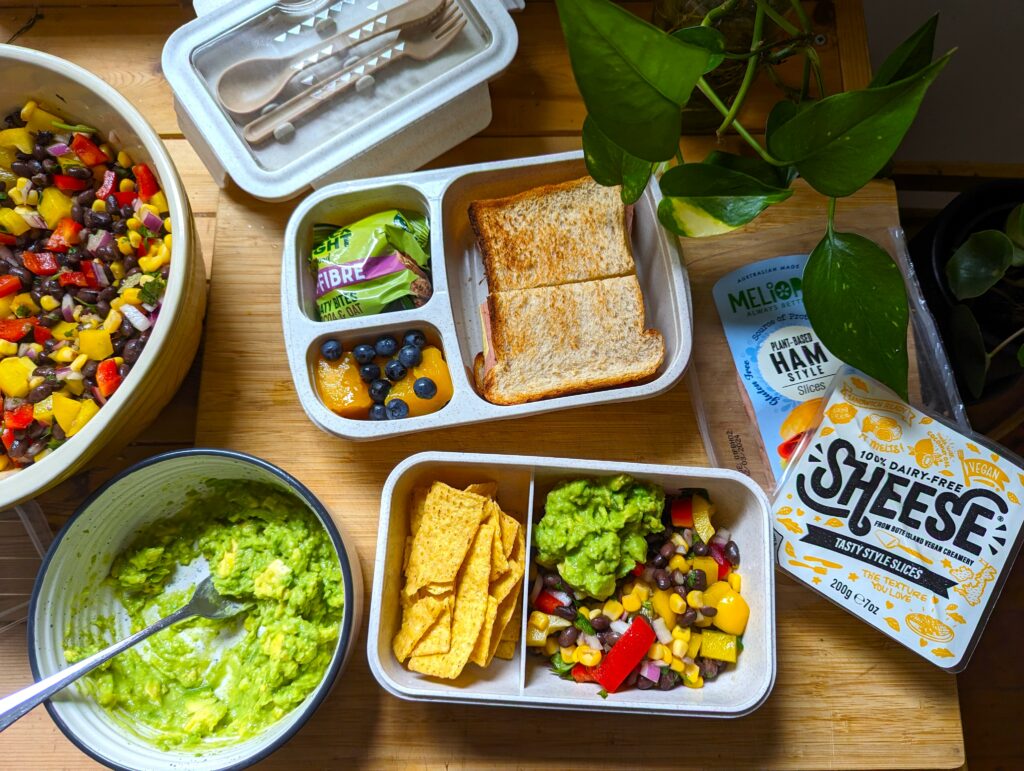 Overhead image of a full bench. Two bento boxes are open and filled with a bean salad, guacamole, tortilla chips, a sandwich, fruit, and some packaged snacks. A full bowl of pre-prepared salad is in shot, along with a bowl of guacamole. Packets of vegan cheddar slices and vegan deli slices sit to the side.