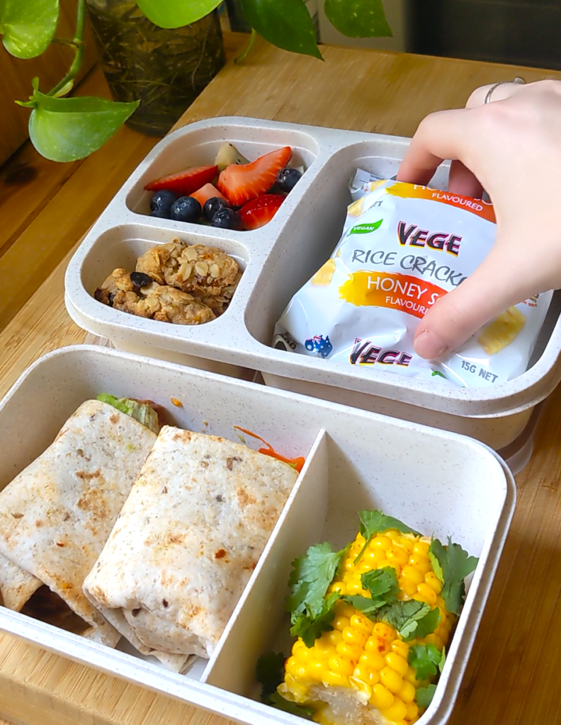 Two bento boxes sit atop a wooden bench, packed with cookies, fruit, wraps, and corn. A hand is adding in a packet of rice crackers.