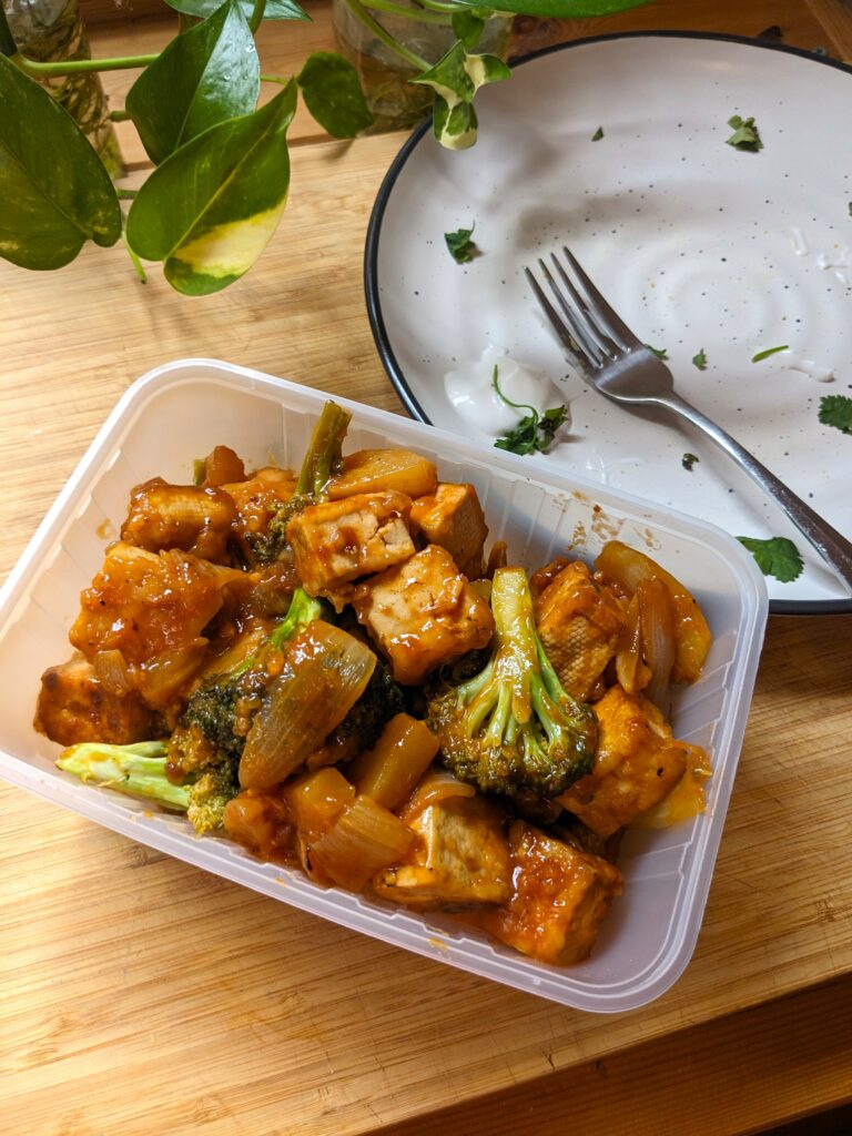 A plastic container of pineapple tofu and broccoli sits on a bench.