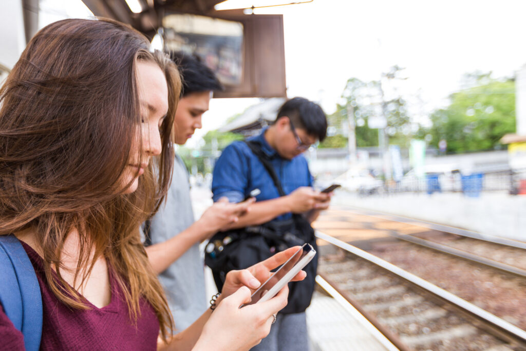 Three people are using their phones while waiting at the train station. 