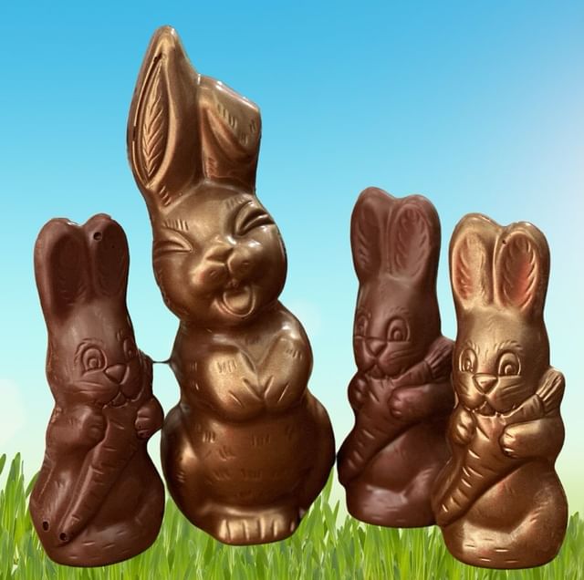 Four chocolate rabbits of different sizes sit in a photoshopped background with green grass and blue skies.