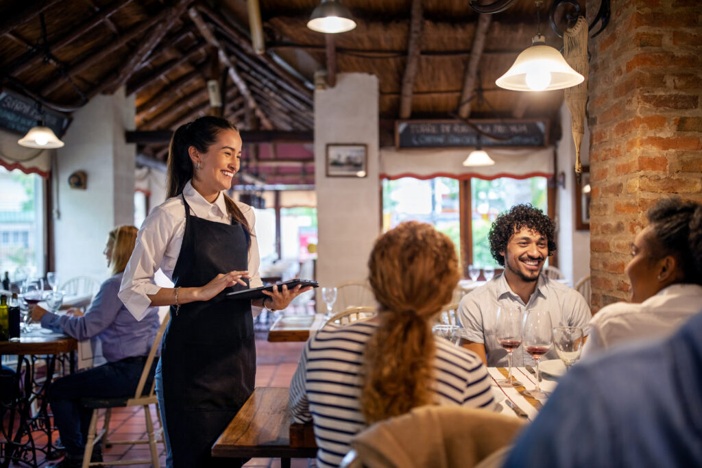 Server at restaurant smiles attentively at a table of customers, holding her tablet.