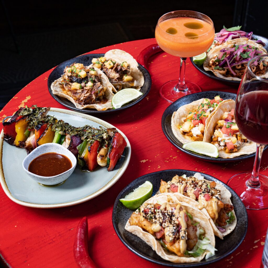 An assortment of Mexican-style dishes are on a red table, with a glass of wine and a colourful orange cocktail to the side.