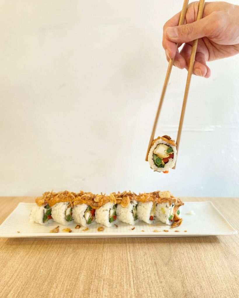 Image of a tray of sushi rolls on a wooden table presented beautifully. A hand is just in shot holding one of the rolls in chopsticks to the front of camera, showing a filling of tofu, capsicum and cucmber.