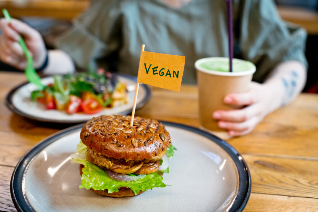 On a wooden cafe table, someone sits eating a salad with a drink, while a burger is in the foreground with flad saying 'vegan'.