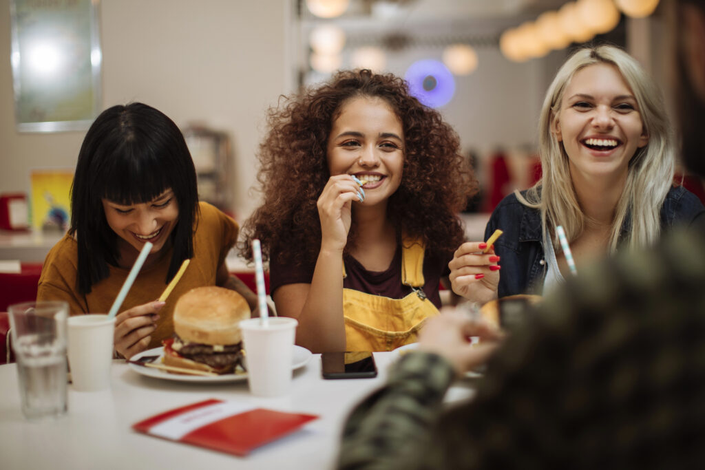 Three woman are eating at a fast food restaurant, sitting opposite another person who they are laughing with.