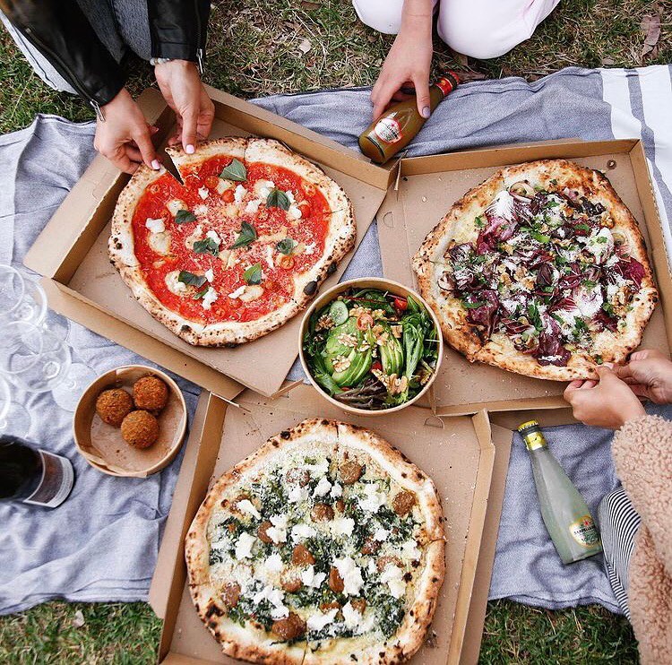 An overhead shot of a picnic rug with three people reaching in to grab food. There are drinks, three large pizzas, a salad, and arancini on display.