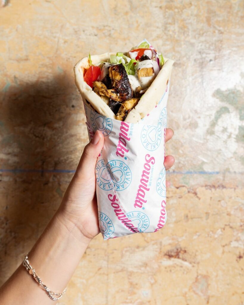 An arm holds up a souvlaki wrapped in an 'I Should Be Souvlaki' wrapper. It's full of salad, condiments, and thick and juicy plant-based meat.