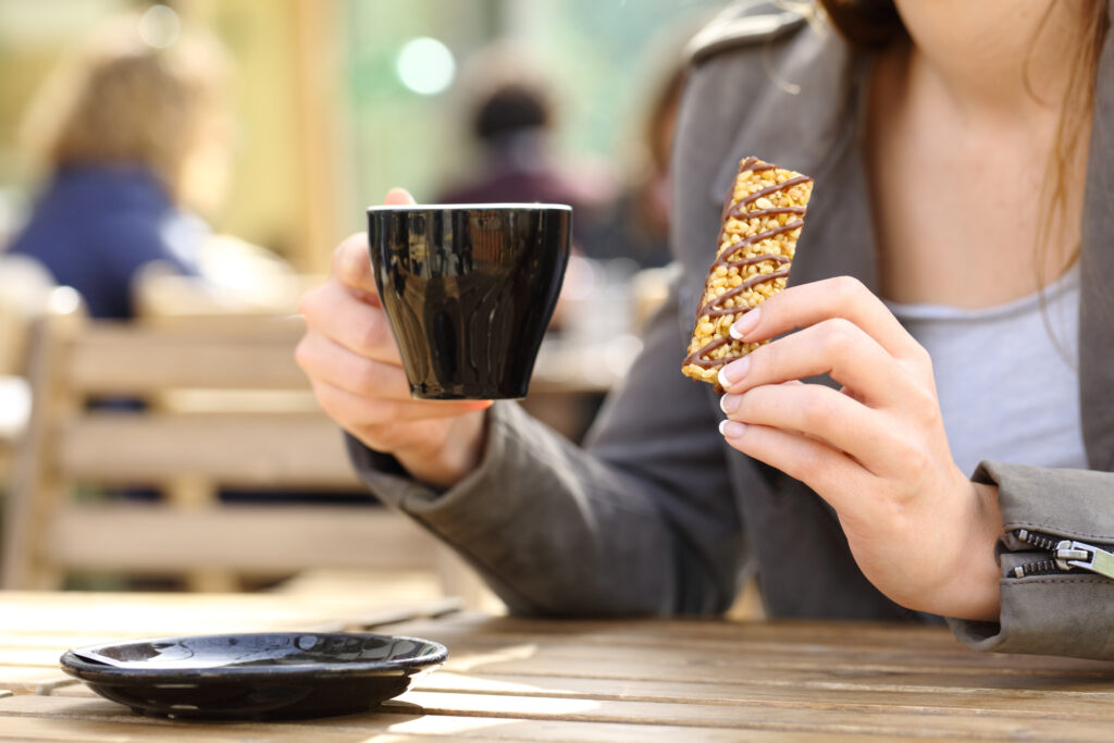 A shot of a woman at a cafe holding a cup in one hand and a muesli bar in the other.