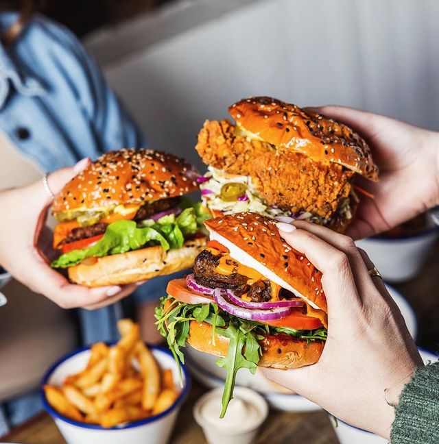 Three hands are 'cheers-ing' with delicious looking vegan burgers. Fries and condiments are visible below on the table.