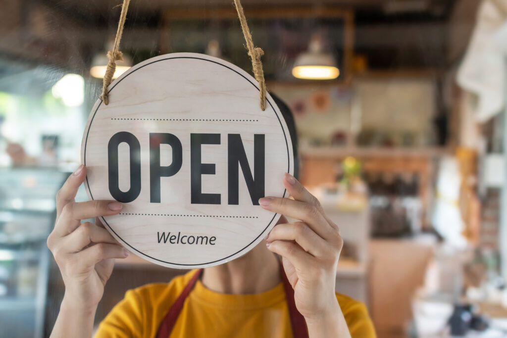 A person is turning the sign on the front door of a cafe to 'open'. Their face is obscured by the sign.