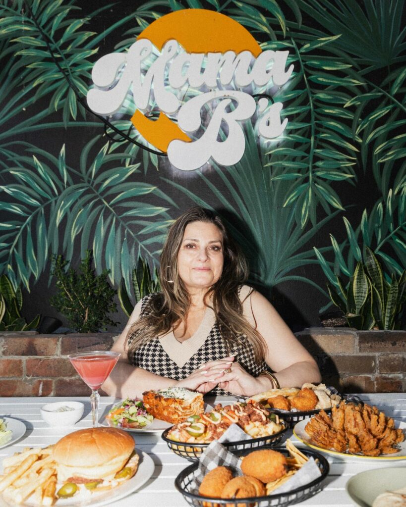 Bhavani Baumann, the publican behind the Chippo, sits at a table against a wall covered with a mural of greenery. She sits under the Mama B's logo, which is the name of the bistro where her vegan creations are served. She has several meals in front of her to showcase, along with a cocktail.