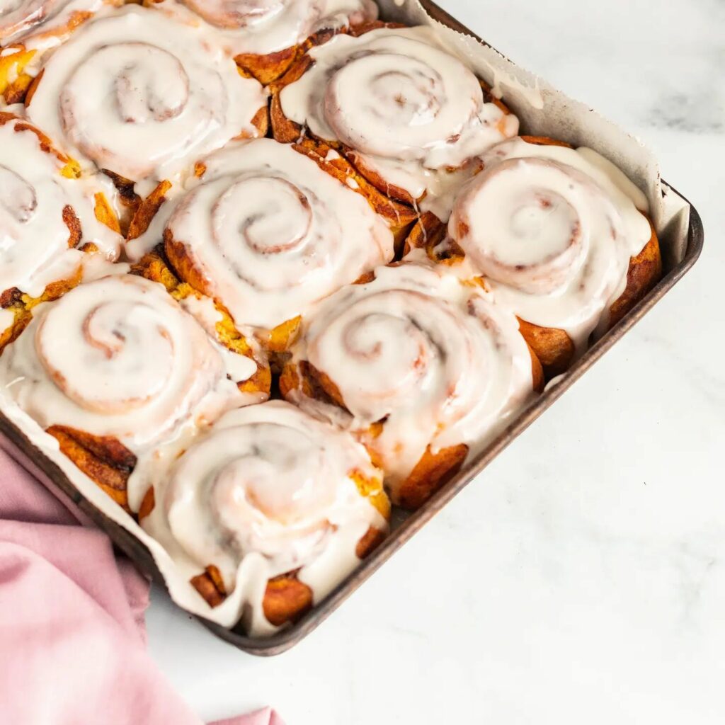 Overhead image of a tray full of iced cinnamon scrolls on a bench. There is a pink tea towel underneath the tray in the corner of the shot.