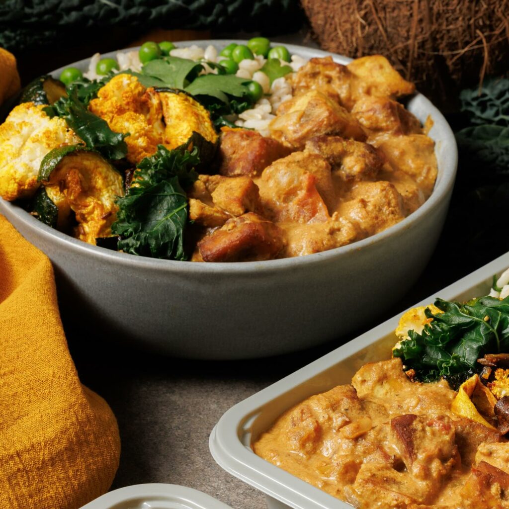 A bowl of butter tofu makhani with rice sits on a table alongside another tray of the dish.