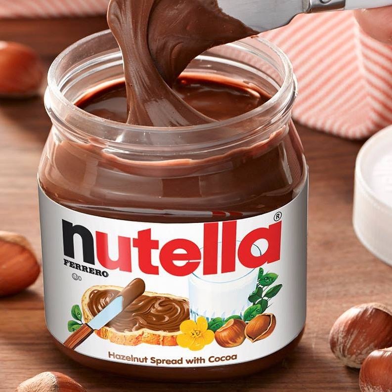 A spatula sits above an open jar of Nutella, covered in the spread. Around it sits full hazelnuts.