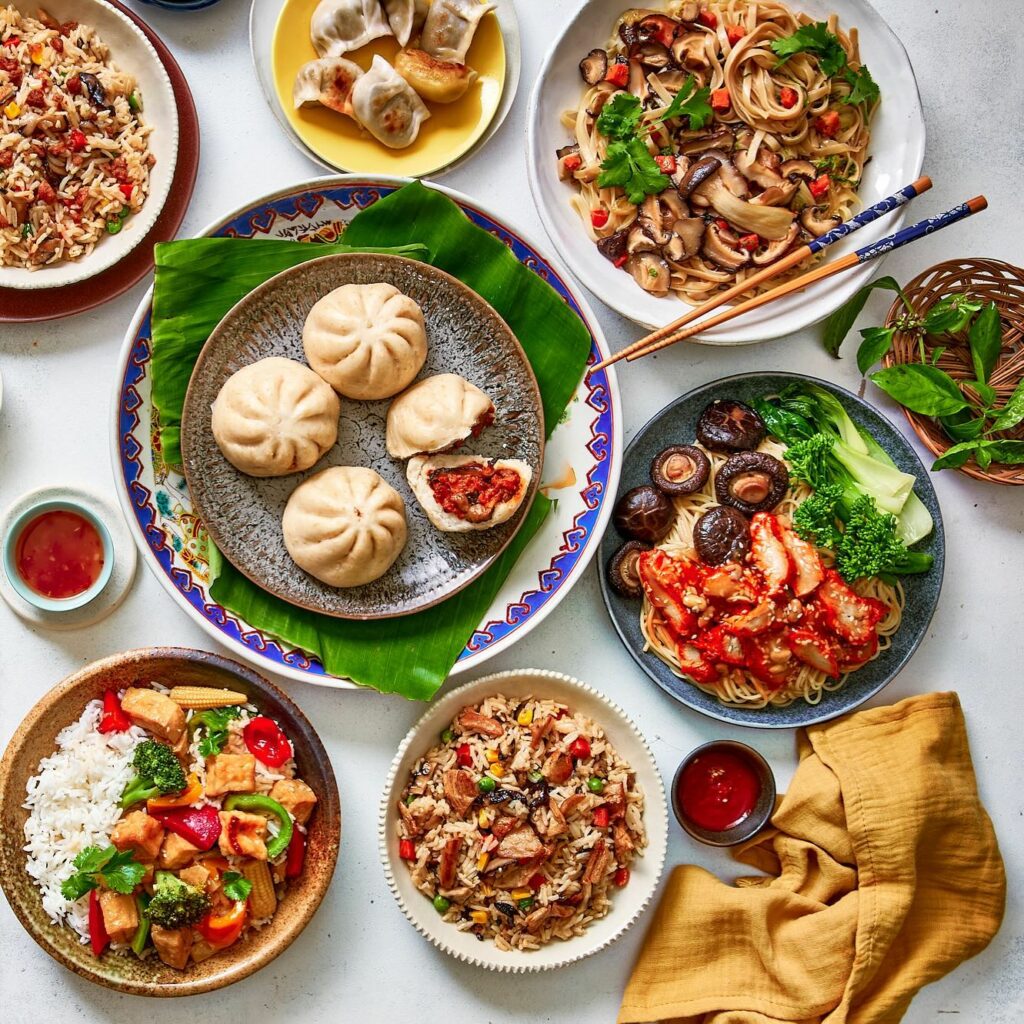 An overhead shot of an array of dishes on a table, including curries, stir-fries, filled buns and gyoza.