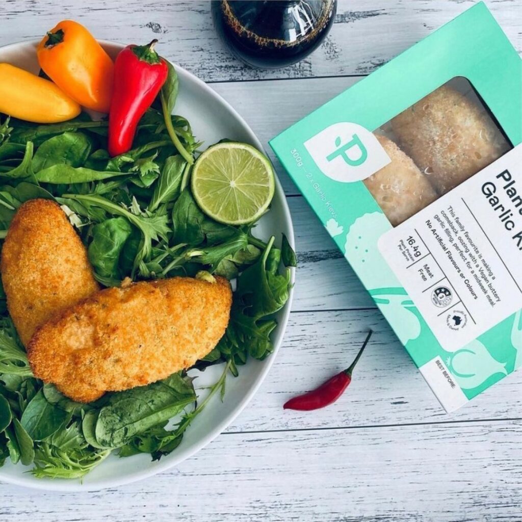 An overhead image of two plant-based garlic Kievs on a bed of spinach. The product box sits next to it revealing it's Plantein brand.